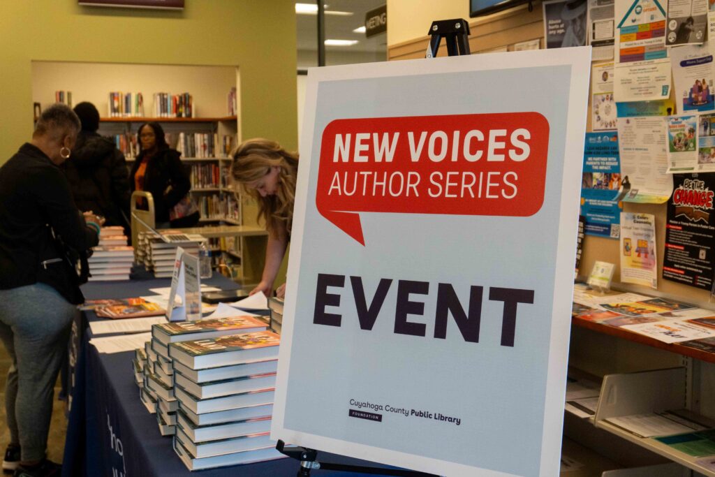 A sign reading "New Voices Author Series Event" stands next to a table full of books.